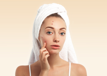 Close up isolated portrait of beautiful young Caucasian woman with blue eyes and problem skin, looking at the camera, pointing at pimple wearing white towel on her head against blue studio background